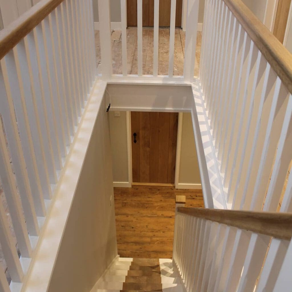 View of a wooden staircase with white railings, leading down to a lower floor. The stairs have a wooden floor, and there's a wooden door visible on the lower level. The overall setting appears to be indoors.