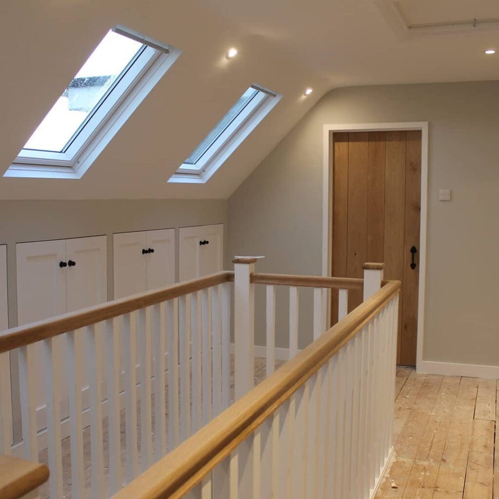 A hallway with wooden flooring and a wooden door. The white railing and paneling contrast with the light green walls. Three skylights are on the angled ceiling, allowing natural light to illuminate the space.