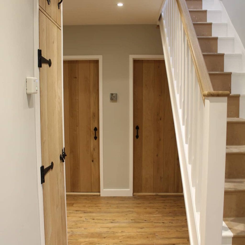 A hallway with wooden flooring and light gray walls features three wooden doors with black handles and hinges. A staircase with wooden steps and white railings is on the right side, leading upwards. The space is well-lit by ceiling lights.