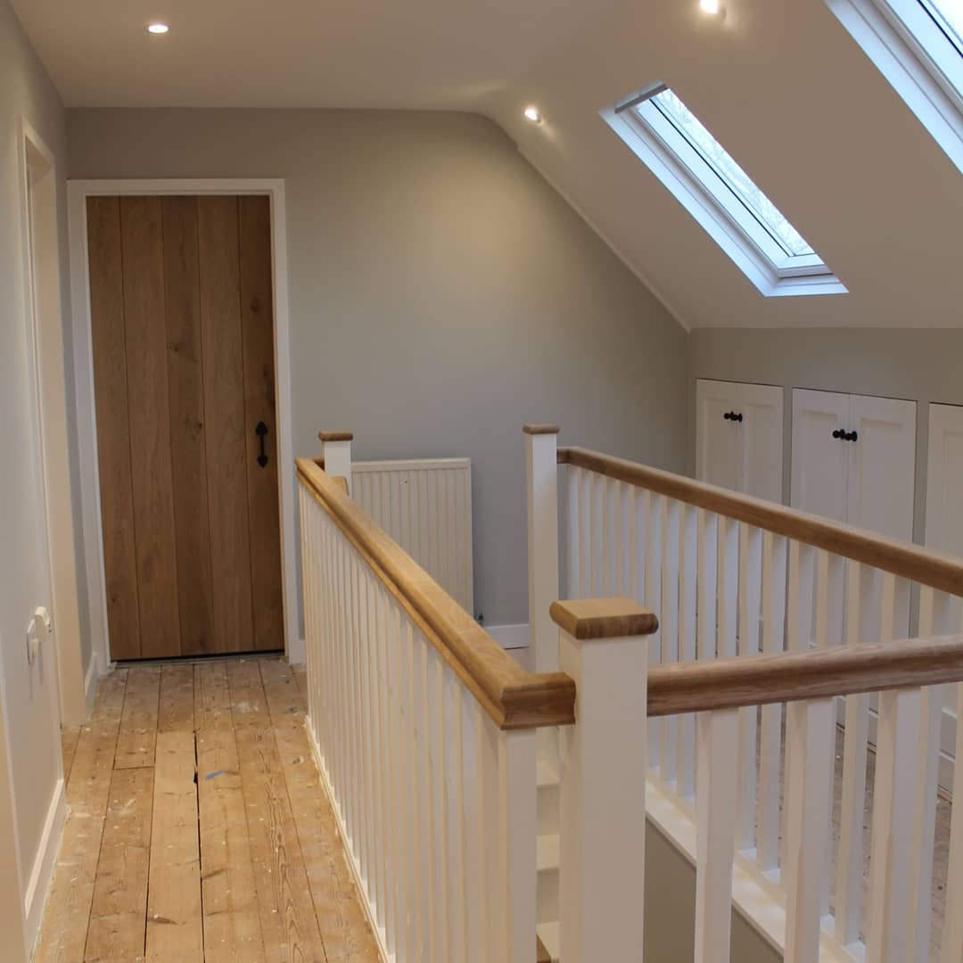 A bright, modern hallway with a wooden floor and white walls. A wooden door is at the end of the hall, and there are two skylights on the sloped ceiling. The hallway features white railings with wooden tops and built-in cabinets on one side.