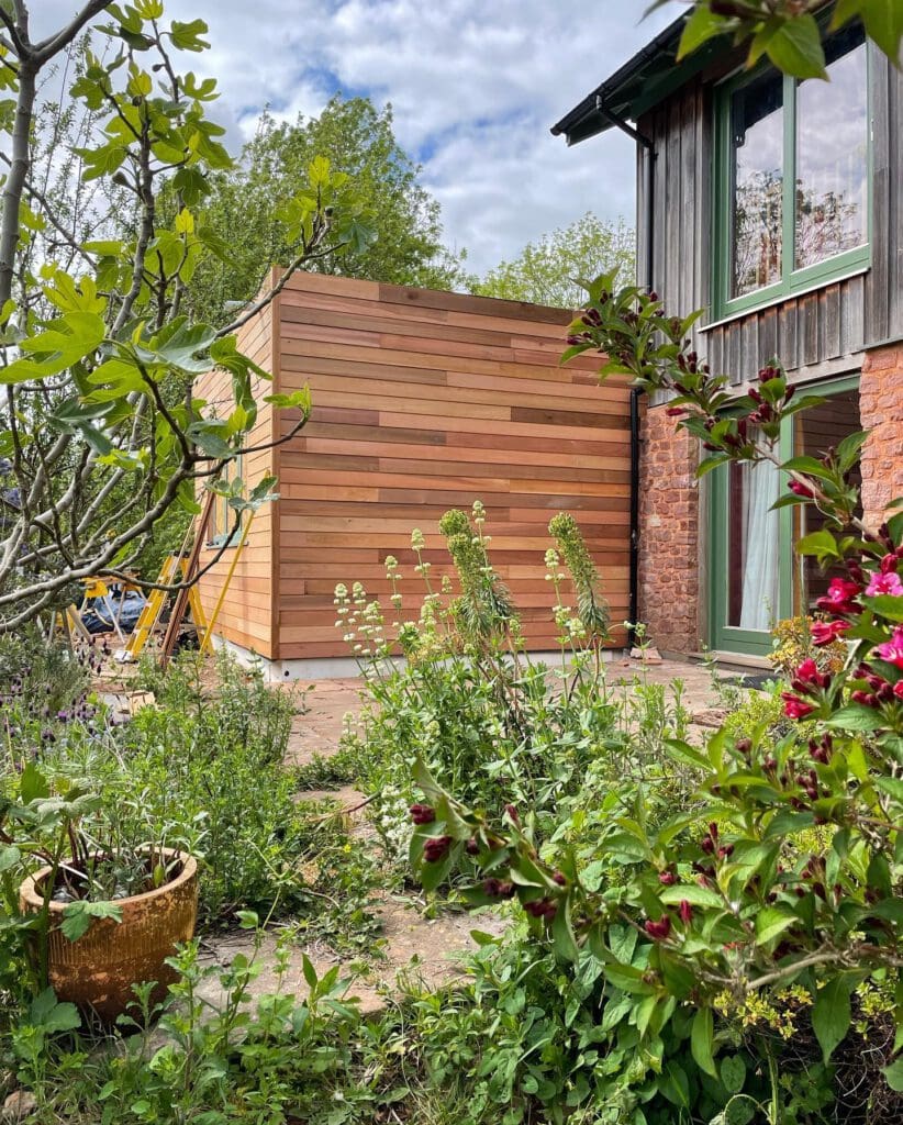 A garden scene with a mix of green plants and flowers in front of a modern house. The house has brick and wooden siding, large windows, and a small patio area. The sky is partly cloudy.