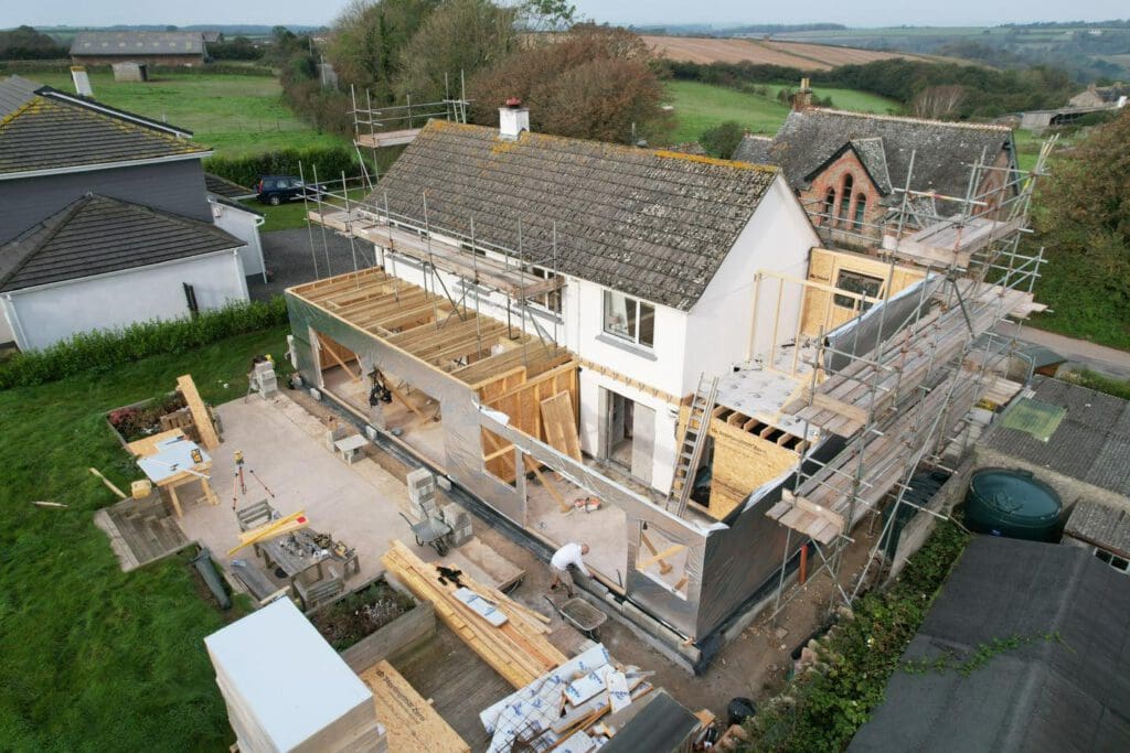 Aerial view of a house under construction, surrounded by scaffolding. The roof and walls are partially completed with visible wooden beams. Workers are present, and there is a grassy area and neighboring buildings around the site.