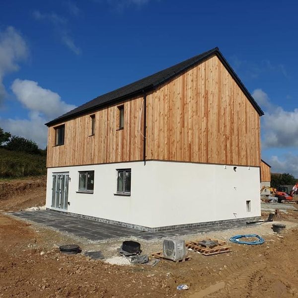 A two-story building under construction with a wooden facade on the upper level and white stucco on the lower. It's set on a plot of earth, with construction materials and tools nearby. The sky is clear and blue.