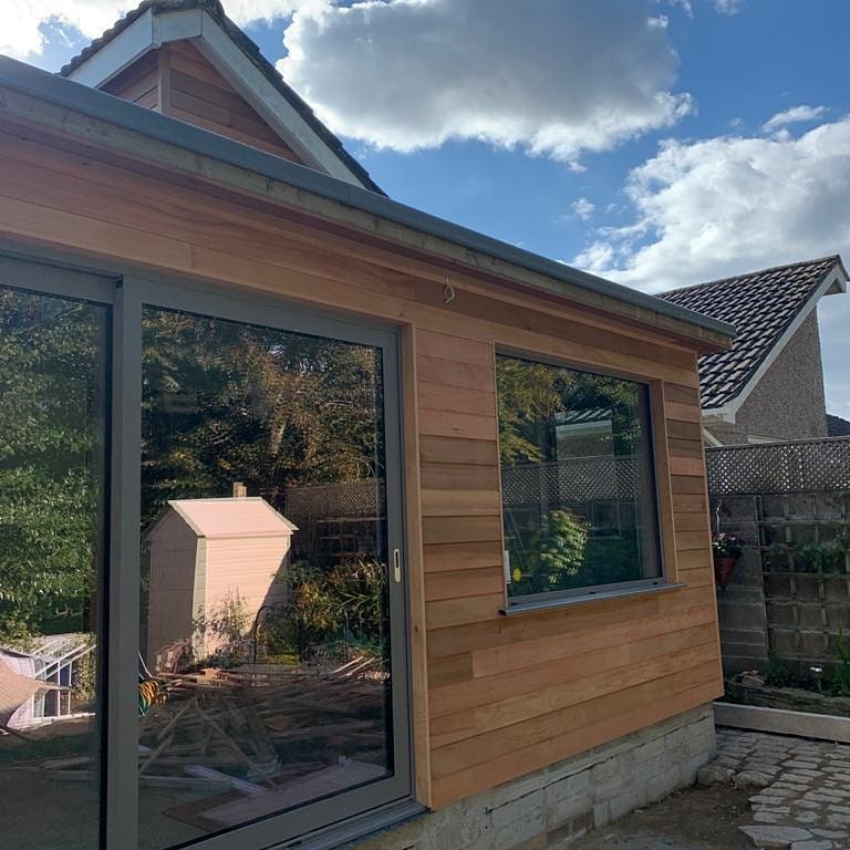 A modern wooden extension with large glass sliding doors on a sunny day. The reflection of trees is visible in the glass. The sky is partly cloudy, and a small white shed and stone pathway are in the surrounding area.