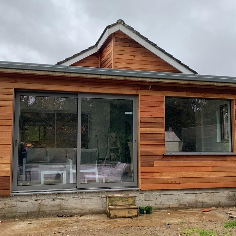 A small, modern house with wood paneling and a large glass sliding door. There are two small potted plants on the ground in front of the door. The sky is overcast.
