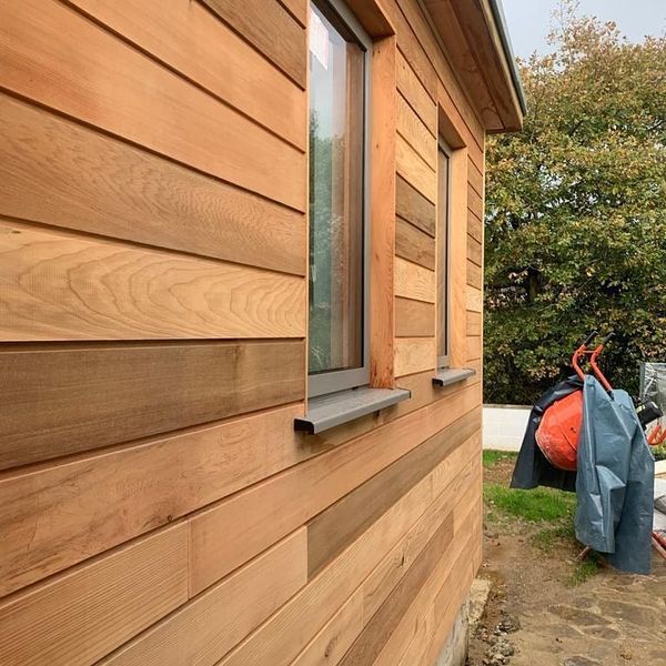 Wooden-clad exterior of a house with horizontal planks. Two windows have gray sills. A wheelbarrow and tarp-covered items are in the background near a tree. The ground is partially paved with stones.