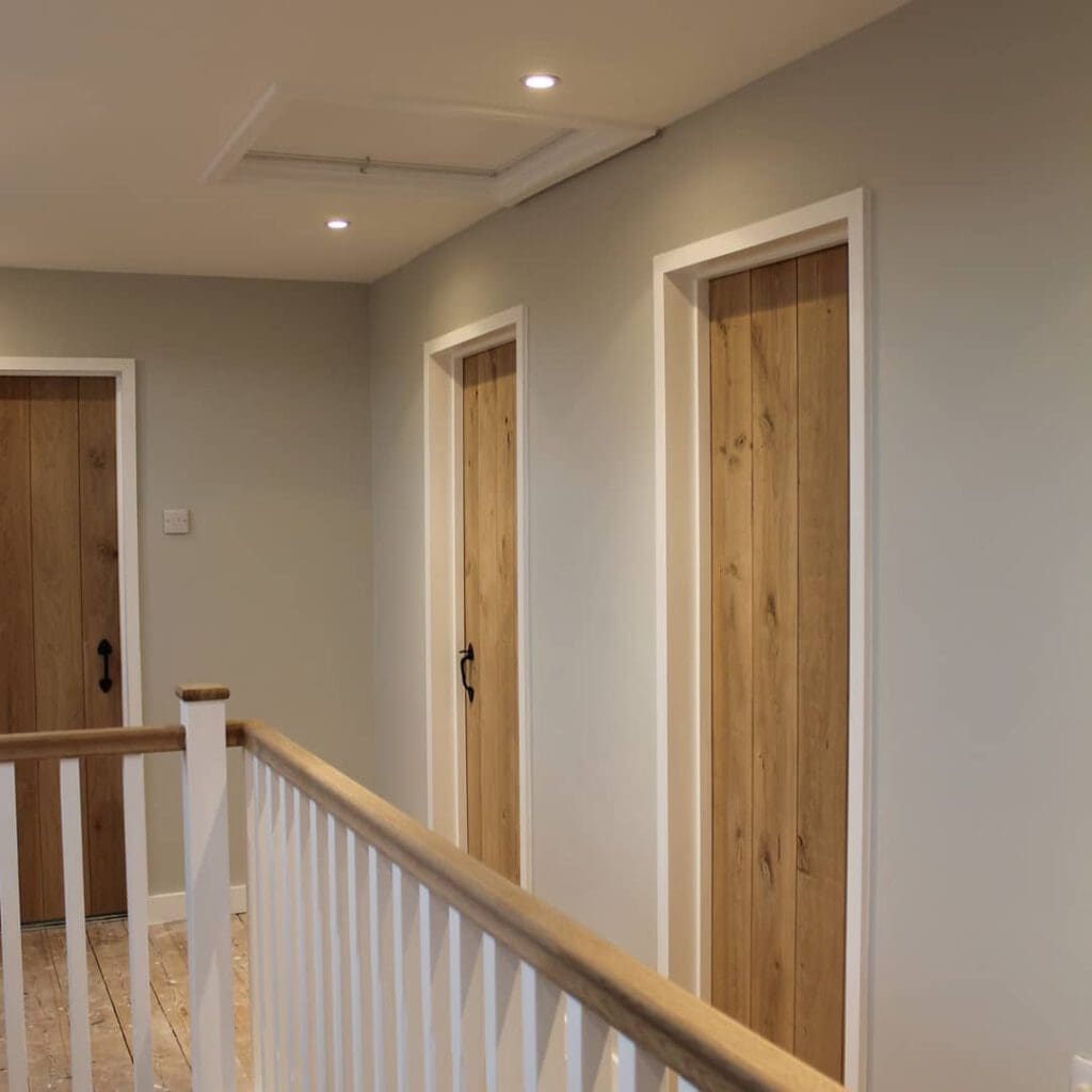 A hallway with wooden doors and white trim. The walls are painted light gray, and the ceiling has recessed lighting. A white railing with a wooden handrail runs along the left side of the image, suggesting a stairway or landing.
