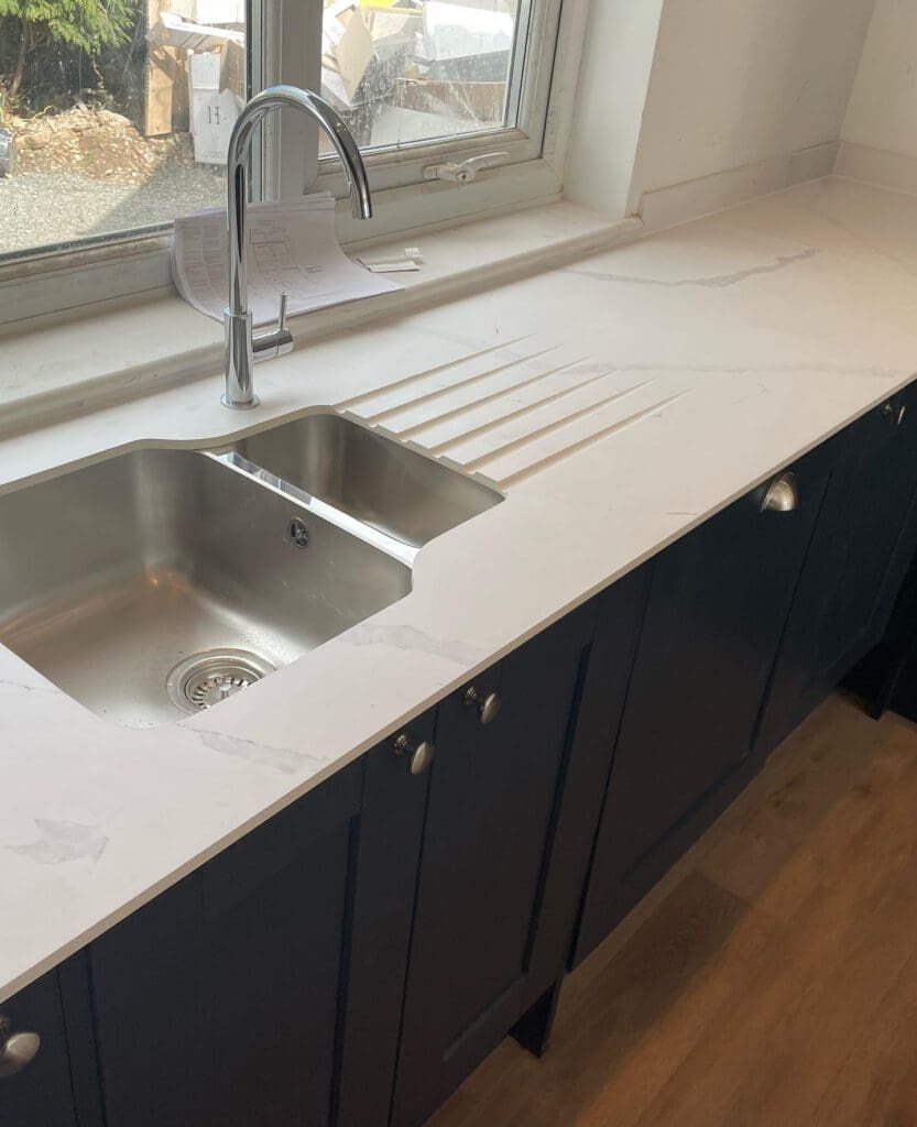 Modern kitchen scene featuring a sleek stainless steel sink with a single faucet, set into a white marble countertop. Black cabinets below add contrast, and a window above offers natural light. A paper rests near the faucet.