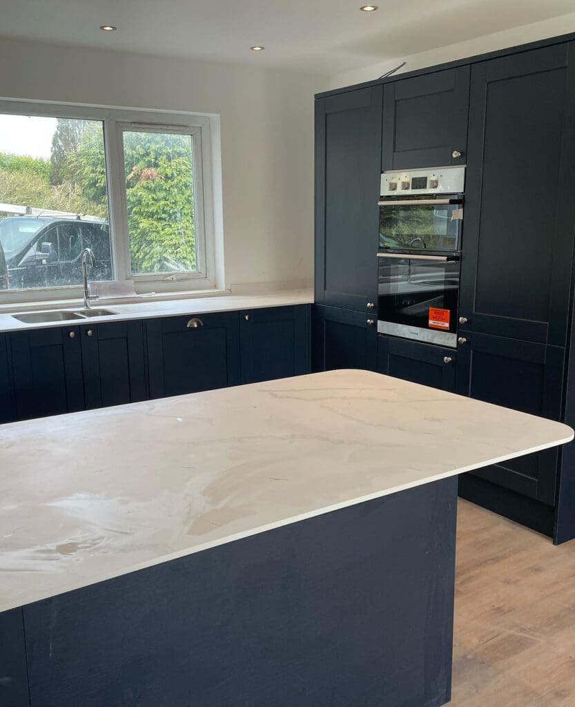 Modern kitchen with dark cabinets, a white marble countertop island, and an integrated oven. A sink is located under a window with a view of greenery outside. Recessed ceiling lights illuminate the space.