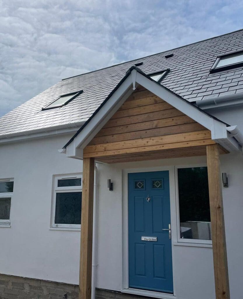 A modern house with white walls and a gray tiled roof. It features a bright blue front door under a covered porch with wooden beams. There are two windows beside the door, and the sky is partly cloudy above.