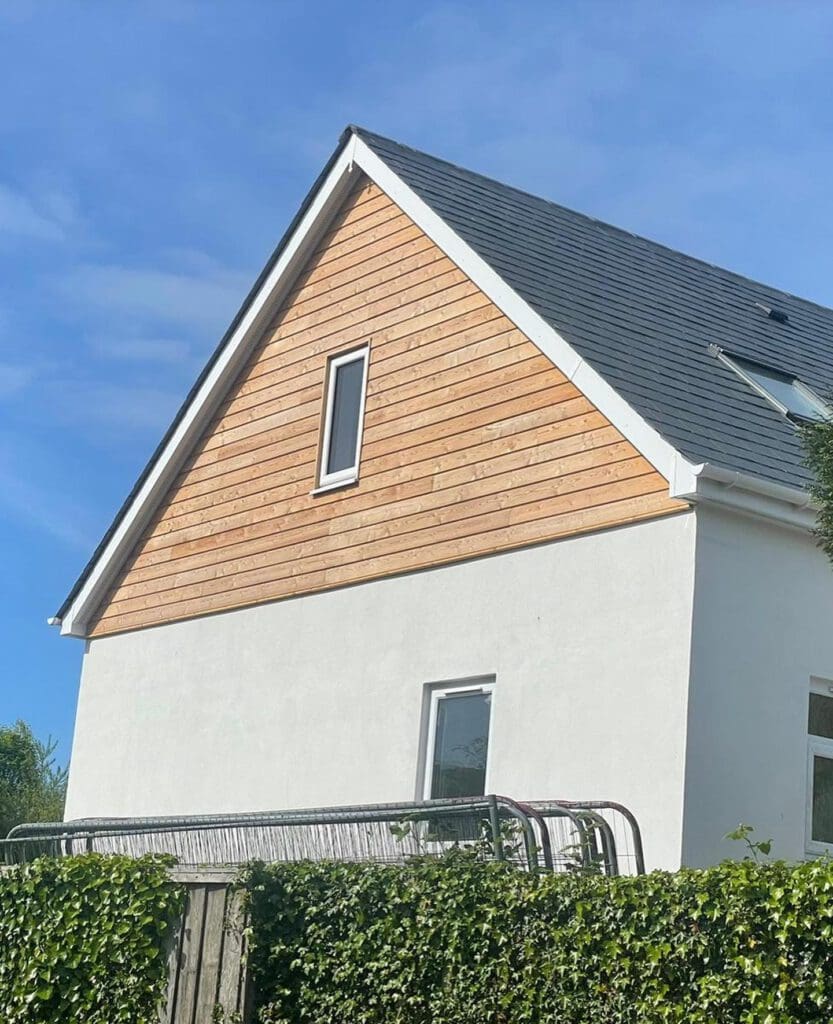 A modern house with a triangular roof, white walls, and horizontal wooden panels under the eaves. A small window is visible near the roof, and a larger one on the ground floor. In the foreground, a hedge and part of a metal gate are visible.