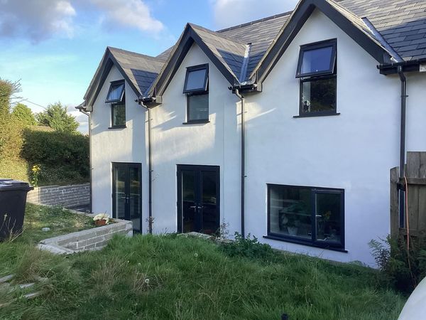 A modern two-story house with a white exterior and black trim. It features multiple triangular dormer windows on the roof. The backyard has grassy areas, a small block planter, and a recycling bin on the left. A wooden fence is on the right.