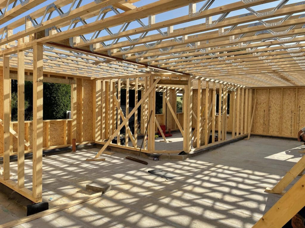 Construction site of a wooden house frame under a clear blue sky. The structure includes upright beams and trusses, with sunlight casting shadows on the floor. There are no external walls, providing a view of the framework's open layout.