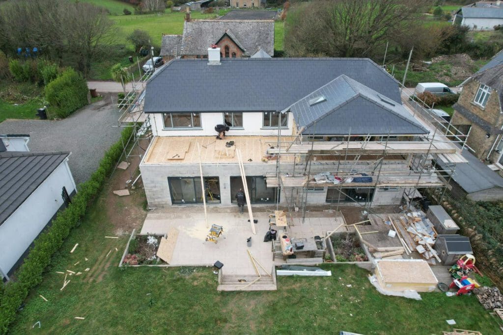 Aerial view of a construction site showing a partially completed house with scaffolding on one side. Workers are on the roof and ground, surrounded by building materials. The yard is cluttered with tools and debris. Nearby houses and greenery are visible.