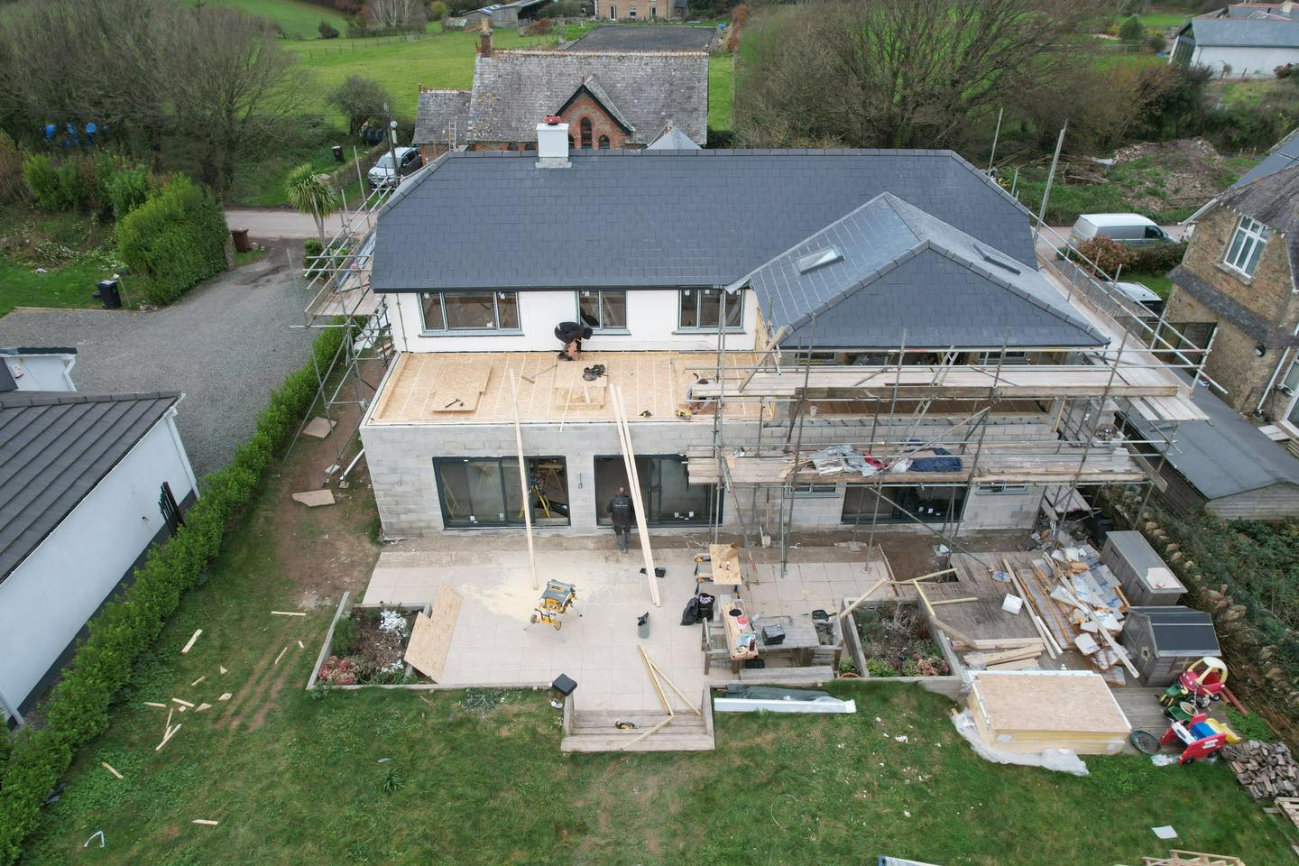 Aerial view of a construction site showing a partially completed house with scaffolding on one side. Workers are on the roof and ground, surrounded by building materials. The yard is cluttered with tools and debris. Nearby houses and greenery are visible.