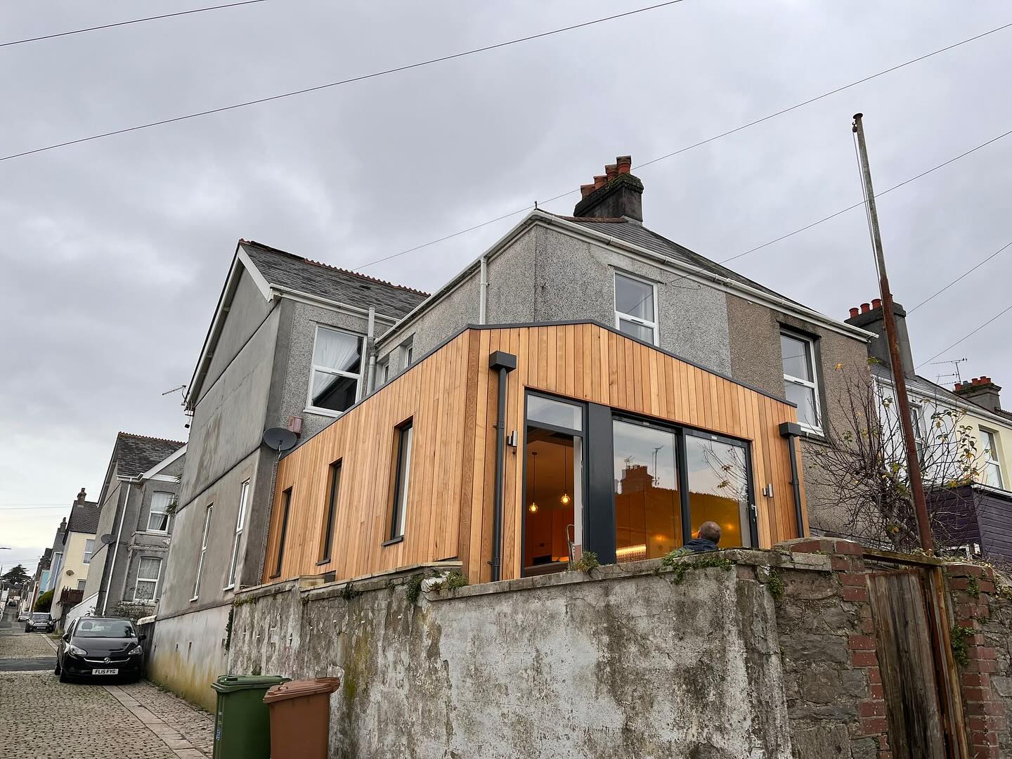 A two-story corner house with a mix of gray and wooden exterior. Large glass doors and windows are present on the wooden section. The sky is overcast, and there’s a nearby parked car and trash bin on the cobblestone street.