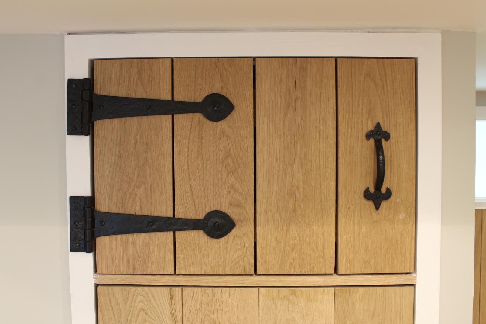 A wooden cupboard door with a vertical plank design, featuring two large black metal hinges on the left and a black metal handle on the right. The door is framed in white.