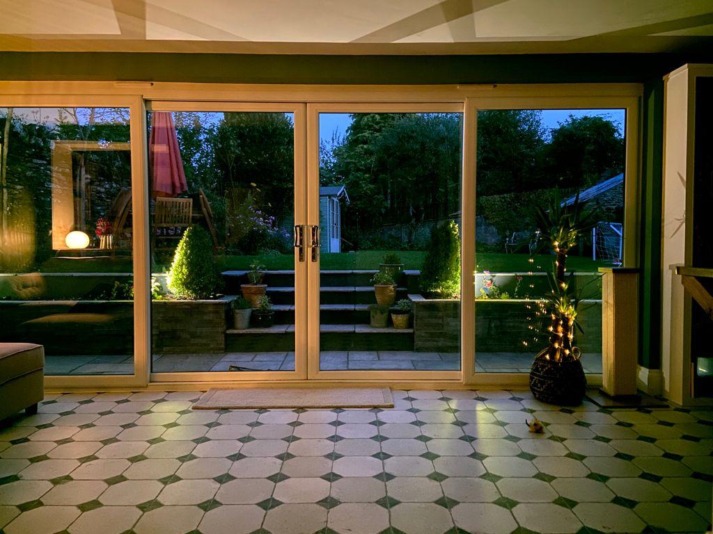 A cozy room with tiled flooring overlooks a garden through large glass doors. Outside, there are plants and steps leading up from a patio. Warm lighting illuminates the plants and surrounds, creating a peaceful evening atmosphere.