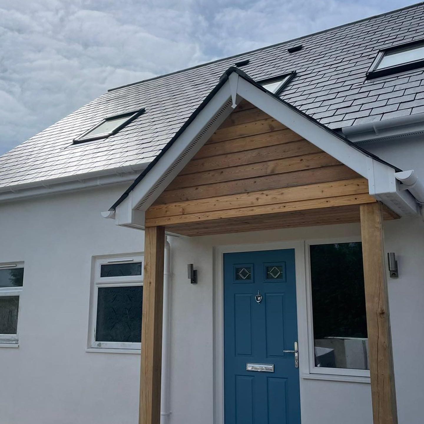 A modern house with white walls and a gray tiled roof. It features a bright blue front door under a covered porch with wooden beams. There are two windows beside the door, and the sky is partly cloudy above.