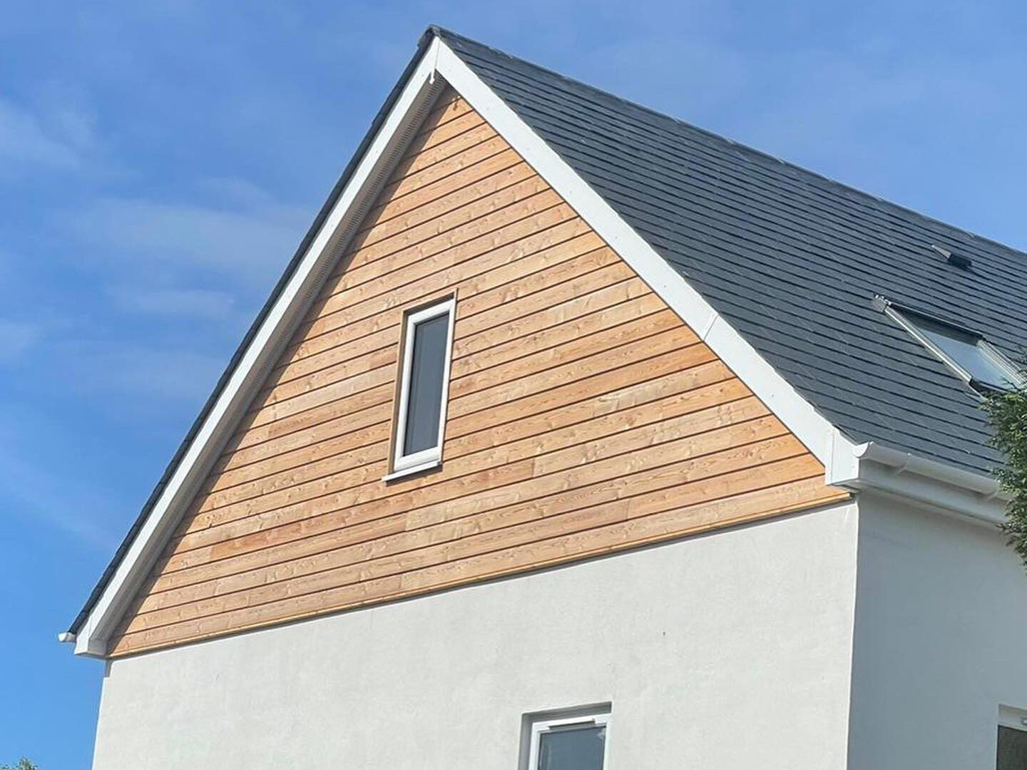 A modern house with a triangular roof, white walls, and horizontal wooden panels under the eaves. A small window is visible near the roof, and a larger one on the ground floor. In the foreground, a hedge and part of a metal gate are visible.